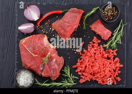 Frisches Rindfleisch forcemeat und Stücke vom Rinderfilet auf schwarzem Schiefer Platte auf schwarzen Holztisch mit Salz, Pfeffer, Chili und frischem Rosmarin, Ansicht von Stockfoto