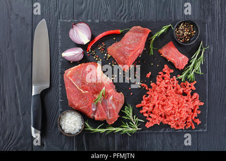 Frisches Rindfleisch forcemeat und Stücke vom Rinderfilet auf schwarzem Schiefer Platte auf schwarzen Holztisch mit Messer, Salz, Pfeffer, Chili und frischem Rosmarin, vie Stockfoto