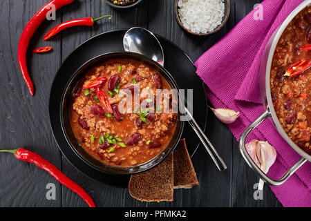 Ein leckeres Chili con carne mit ganzen Red Hot Chili, Bohnen, Tomaten serviert in schwarz Schüssel, einen Topf und Zutaten auf Hintergrund, authentische r Stockfoto