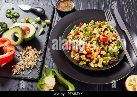 In der Nähe des mexikanischen Stil Salat mit Avocado, schwarze Bohnen, Maiskörner, Buchweizen Sprossen, gewürfelt Pfeffer auf schwarzem Teller serviert mit Zutaten aus sl Stockfoto