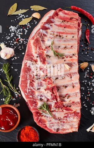 Rack von rohem Schweinefleisch Spare Ribs gewürzt mit Gewürzen auf schwarzem Schiefer Fach mit Paprika, Knoblauch, Nelken, Lorbeerblätter im Hintergrund, vertikale Ansicht von oben, Stockfoto