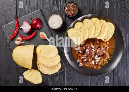 Köstliche hausgemachte heiße Traditionelle tschechische Rindergulasch mit Gedünstetem tschechische Knödel oder knodel auf schwarze Platte auf hölzernen Tisch serviert mit Zutaten Stockfoto