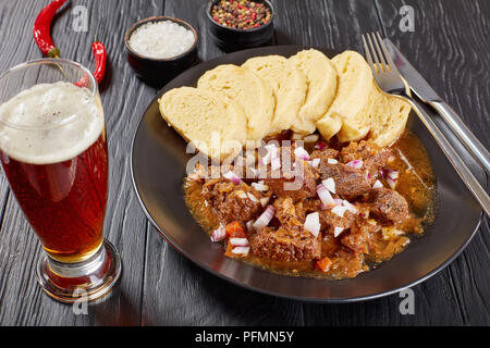 Köstliche hausgemachte heiße Traditionelle tschechische Rindergulasch mit Gedünstetem tschechische Knödel oder Knedliky auf schwarze Platte auf hölzernen Tisch serviert mit Glas bee Stockfoto