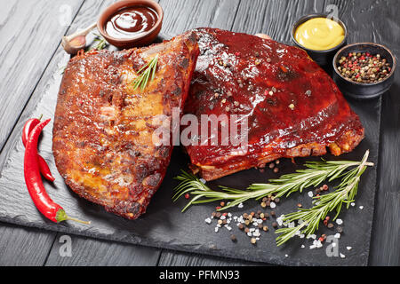 Close-up lecker saftig Hausgemachten geräucherten Grill Schweinerippchen auf schwarzen Stein Platte mit Gewürzen, frischem Rosmarin, Senf und BBQ Sauce, Ansicht von Stockfoto
