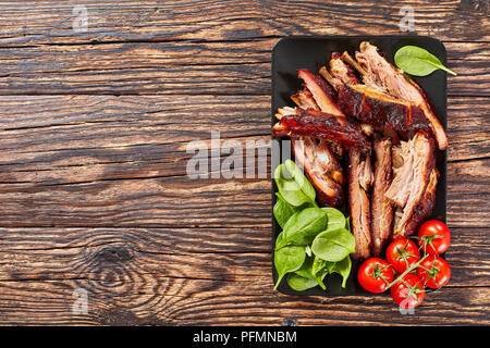 Köstliche geschnitten BBQ Ribs mit frischen grünen Spinat ad Kirschtomaten auf schwarzem Schiefer Fach auf Holz- Tabelle, Ansicht von oben Stockfoto