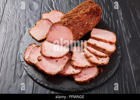 In der Nähe von köstlichen geräucherten Schinken in Scheiben geschnitten, gewürzt mit gemahlene Gewürze auf schwarzem Schiefer rundes Tablett auf schwarzem Holz- Tabelle, Ansicht von oben Stockfoto