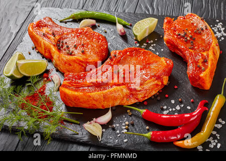 Raw Schweinekoteletts vorbereitet zu kochen, mit Gewürzen und roten sriracha Soße auf schwarzem Schiefer Fach mit Chili Pfeffer, Salz, Kalk Schichten und Gewürzen mariniert, vie Stockfoto