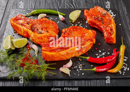 In der Nähe von roh marinierte Schweinekoteletts vorbereitet zu kochen, mariniert mit Gewürzen und roten sriracha Soße auf schwarzem Schiefer Fach mit Chili Pfeffer, Salz, Kalk Stockfoto