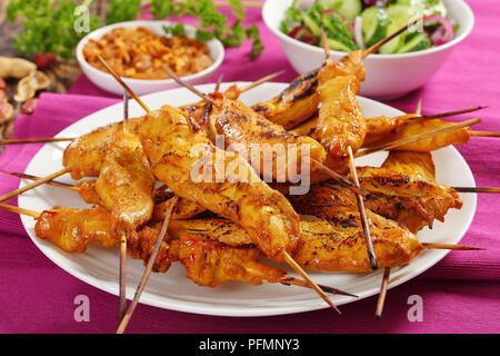 Close-up Chicken Satay auf Spieße auf einem weißen Teller auf rustikalen Holztisch mit Erdnusssoße und Gurke Zwiebel Salat serviert, authentische Rezept, Anzeigen Stockfoto