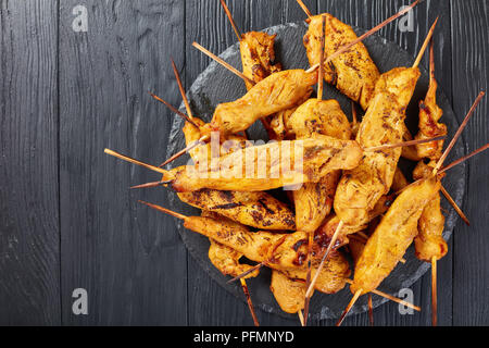 Chicken Satay auf Spieße auf einem schwarzen Stein Fach auf einem Holztisch, vertikale Ansicht von oben, in der Nähe Stockfoto