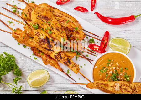 Gegrillte spicy Chicken Satay auf Spieße auf einem weißen rechteckigen Platte mit Erdnusssoße. rote Chilischoten, Kalk Schichten und Petersilie im Hintergrund, Anzeigen Stockfoto