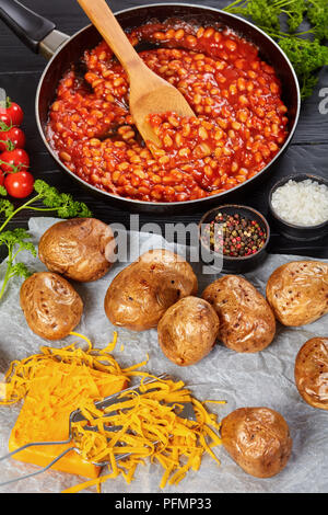 Gebackene Kartoffel oder Pellkartoffeln mit Goldgelb knusprige Haut auf einem Papier mit Cheddar Käse. gebackene Bohnen mit Tomatensauce in einer Pfanne im Hintergrund, Stockfoto