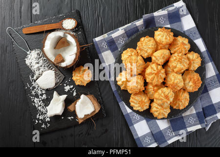 Leckere hausgemachte coconut Makronen Cookies auf einer Platte auf einem schwarzen Holztisch mit Zutaten auf ein Schneidbrett, horizontale Ansicht von oben, flach La Stockfoto