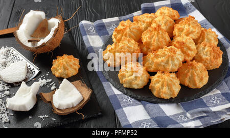 Leckere hausgemachte coconut Makronen Cookies auf einer Platte auf einem schwarzen Holztisch mit Zutaten auf ein Schneidbrett, horizontale Ansicht von oben, in der Nähe - u Stockfoto