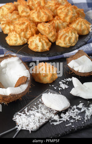 Leckere hausgemachte coconut Makronen Cookies auf einer Steinplatte auf einem schwarzen Holztisch mit Zutaten im Vordergrund, Ansicht von oben Stockfoto