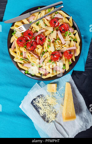 Pasta penne, Schinken, grüne Erbsen herzhaften Salat mit Parmesan bestreut, gekrönt mit gegrillten Tomaten auf schwarzem Teller mit Stockfoto
