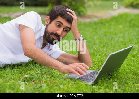 Junger Mann bei der Arbeit auf seinem Laptop beim Liegen auf dem Rasen des Parks. gelangweilter Gesichtsausdruck. Stockfoto