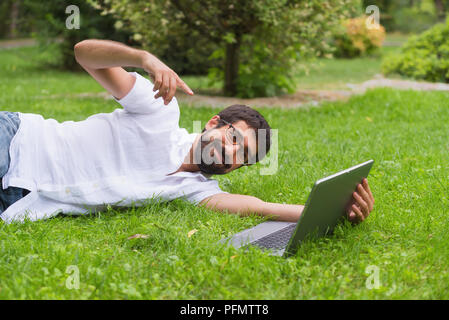 Junger Mann bei der Arbeit auf seinem Laptop beim Liegen auf dem Rasen des Parks. Happy Ausdruck, Begriff der Freiheit. Stockfoto