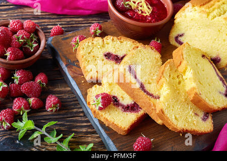 Nahaufnahme der Zitrone Pound Cake mit himbeermarmelade Füllen auf einem Schneidebrett einen alten Holztisch mit frischen Beeren, dunklen rustikalen Stil, die französische Küche, Stockfoto