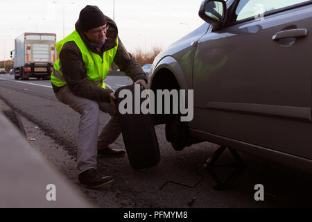 Mann mit hoher Sichtbarkeit jacke Änderungen ein Rad mit Reifen zu einem Auto auf dem Seitenstreifen einer Autobahn gestoppt Stockfoto