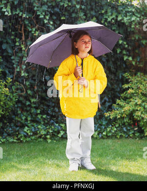 Ständigen Mädchen in gelb Regenjacke hält geöffnete grau Sonnenschirm im Garten, Vorderansicht. Stockfoto