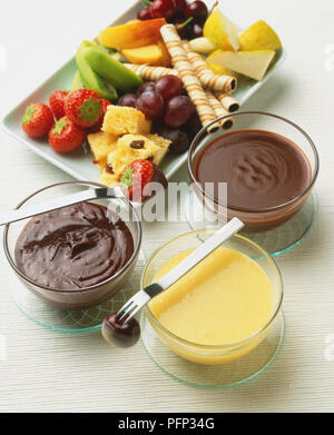 Dunkle Schokolade, weiße Schokolade und chocolate fudge Fondues in Glasschalen, Stücke von frischem Obst, Kuchen und Kekse in Weiß Teller hinter, Ansicht von oben. Stockfoto