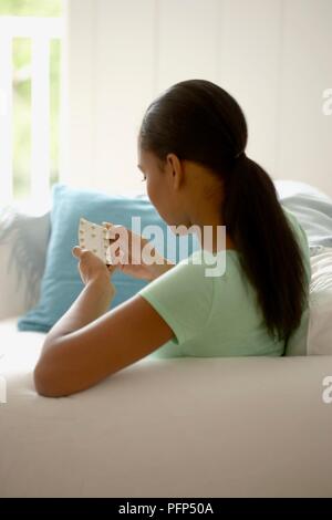 Frau mit schwarzen Pferdeschwanz sitzen auf einem Sofa holding Blister in den Händen, Rückansicht Stockfoto