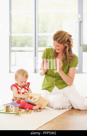 Baby Mädchen sitzen auf dem Boden spielen mit Holzspielzeug, Frau neben ihr sitzt Händeklatschen Stockfoto