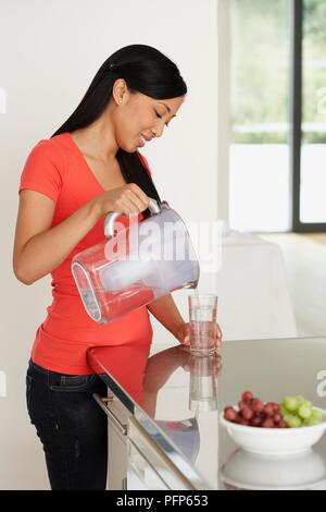 Schwangere Frau gießen Glas Wasser aus dem Filter, Kanne, Schale mit Obst in der Nähe Stockfoto