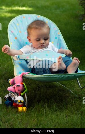 Baby Junge sitzt auf einem Stuhl in einem Garten geschnallt, Spielzeug narby Stockfoto