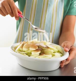 Mädchen mit Löffel Zucker streuen auf Zimt und geschnittene rohe Äpfel in Weiß Auflaufform Stockfoto