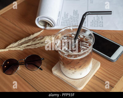 Eis Kaffee im Becher Tasse mit schwarzem Rohr, Handy, Sonnenbrille und Papier auf Holz- Tabelle im Vintage Style Cafe. Kaffee einzigartiges Konzept. Stockfoto