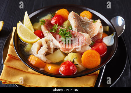 Herzhafte Fischsuppe - Griechische psarosoupa mit Zitronenscheiben in einer schwarzen Schüssel serviert auf einem Holztisch mit Serviette und Löffel, Ansicht von oben, close-up Stockfoto