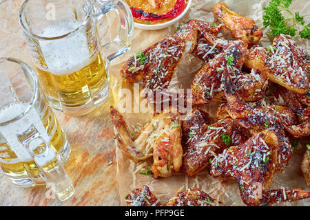Nahaufnahme der knusprig gebackene Chicken Wings mit geriebenem Parmesan bestreut auf einem Papier mit süßen würzigen Pfirsich Salsa und Glas Schalen aus geschäumtem Bier auf Stockfoto