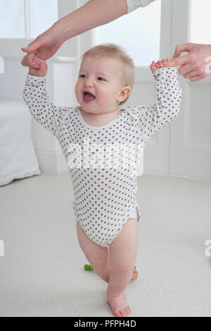 Baby Boy von den Händen seiner Mutter unterstützte, als er lernt, auf dem Teppich zu laufen Stockfoto
