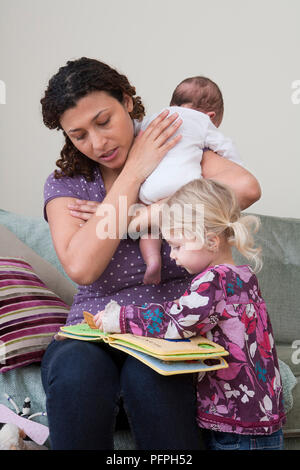 Mutter auf dem Sofa Holding Baby gegen Ihre Schulter, während Jungen Mädchen spielt mit einem Bilderbuch auf Ihrem Schoß sitzen Stockfoto