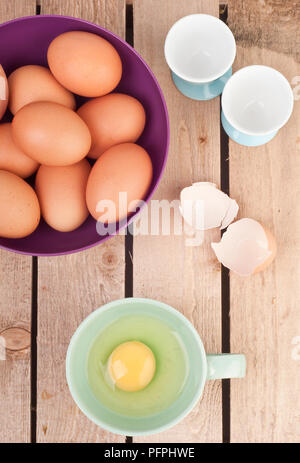 9 Hühnereier in lila Schüssel auf Picknick Tisch neben frischen, rohen Ei in Schale, zerbrochene Eierschale und zwei leere Eierbecher, Ansicht von oben Stockfoto