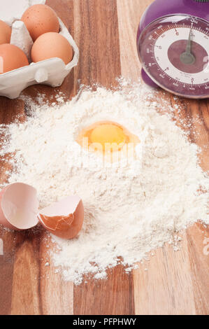 Rohes Ei in der Mitte des Haufens von weißem Mehl auf Holztisch mit Box von Eiern, zerbrochene Eierschale- und Küchenwaagen, close-up Stockfoto