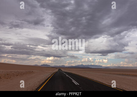 Endlose gerade Teerstrasse mit blauen Berge am Horizont im roten Sand der Wüste mit dramatischen cloudscape gerade vor einem Sandsturm, Namib Naukluft Nati Stockfoto