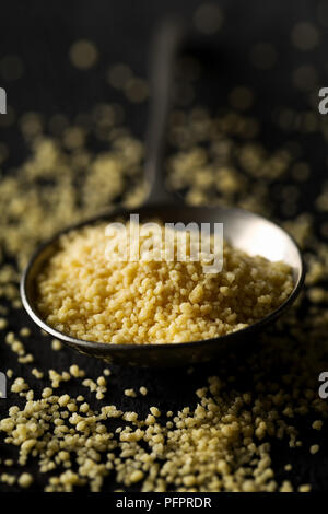 Haufen rohe, ungekochte Couscous in Metalllöffel auf dunklen Holztisch Hintergrund mit selektiven Fokus Stockfoto