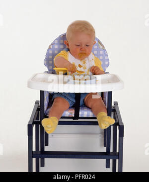 Baby Boy lernen zu füttern selbst sitzen in Kinderhochstuhl Stockfoto