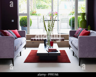 Vase mit Blumen und Bücher über Kaffee Tisch zwischen passende Sofas und Kissen in der Lounge hell durch die französischen Fenster leuchtet Stockfoto