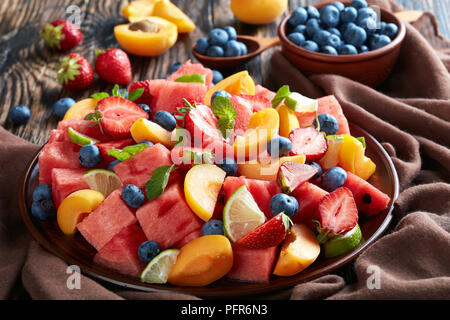 Sweet melone Pfirsich und Berry-Salat mit Minze und Limette Keile auf einem Ton Platte auf einem dunklen Küche Holztisch mit Zutaten, Holzlöffel und nap Stockfoto