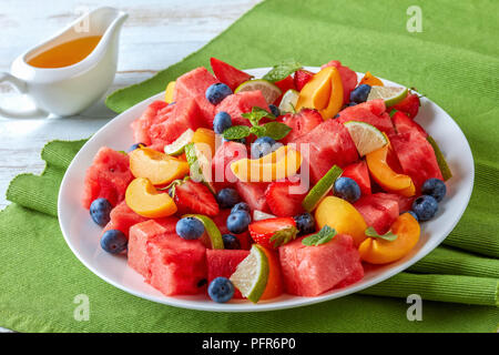 Nahaufnahme der Wassermelone Pfirsich und Berry-Salat mit Minze und Limette Keile auf einem weißen Teller auf einem Holztisch mit süßen Orangen Dressing im Hintergrund Stockfoto