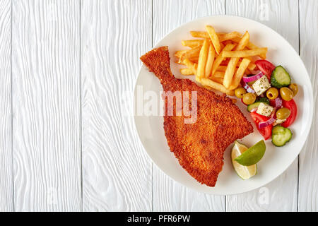 Gebratene Flunder paniert serviert mit frischem Gemüse, Feta, Oliven griechischen Salat und Pommes frites auf einem weißen Teller auf einen hölzernen Tisch, Blick von ab Stockfoto