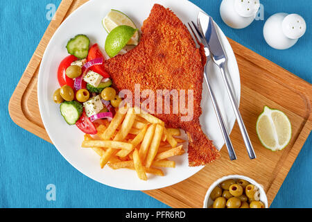 Knusprig gebratene Flunder paniert serviert mit frischem Gemüse, Feta, Oliven griechischen Salat und Pommes frites auf eine weiße Platte mit silbernen Gabel und kni Stockfoto