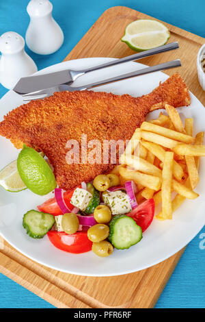 Knusprig gebratener Fisch paniert, mit frischem Gemüse, Feta, Oliven griechischen Salat und Pommes frites auf einem weißen Teller mit Messer und Gabel Stockfoto