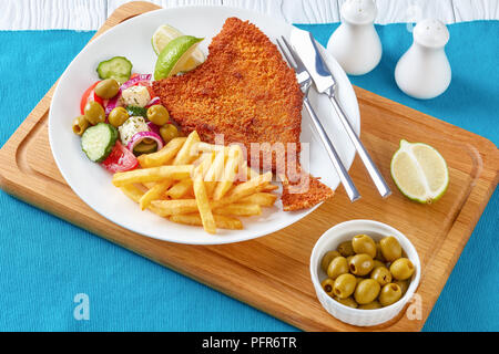 Pan-Fried Flunder mit griechischen Salat und Pommes frites serviert auf einem weißen Teller mit Messer und Gabel auf einem Schneidebrett, Ansicht von oben, close-up Stockfoto