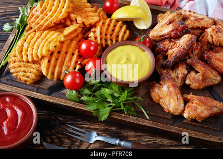 Nahaufnahme der Fried Chicken Wings, knusprige Pommes frites Potato Kreuz und Quer auf einem Ton Platten auf einem Holztisch mit Senf und Tomatensauce eintauchen, Ansicht von eine Stockfoto