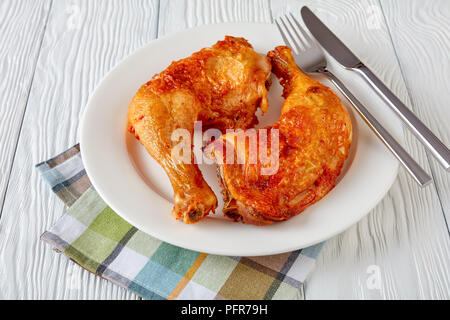 Close-up lecker saftig gebratene Hähnchenschenkel mit goldenen Haut Kruste serviert auf einem weißen Teller mit Messer und Gabel auf einem Holztisch, horizontale Ansicht f Stockfoto